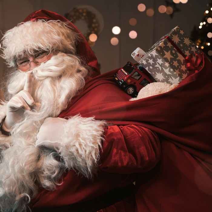 Santa carries a red sack filled with toys and places his finger over his mouth in a "Shhhhh!" gesture.