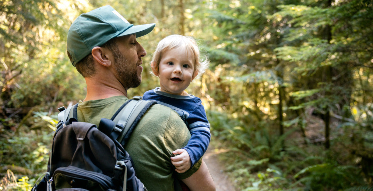 A man wearing a hiking backpack carries his toddler son in his arms while heading down a forested trail. 