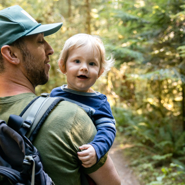 A man wearing a hiking backpack carries his toddler son in his arms while heading down a forested trail. 
