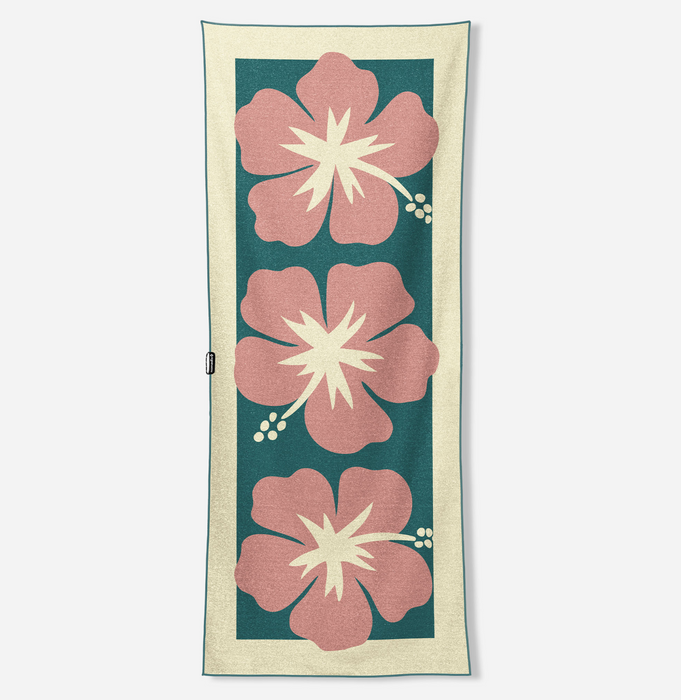 A rectangular woven textile with a cream border and three large pink hibiscus flowers on a dark teal background, shown flat against a light gray studio backdrop.