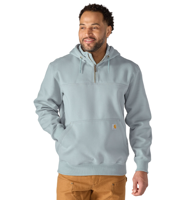 A man wearing a light blue-gray pullover hoodie with a half-zip neckline, drawstring hood, and a small Carhartt logo patch on the front pocket, paired with brown pants against a plain white background.
