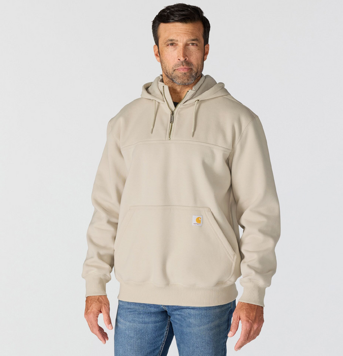 A man wearing a light beige Carhartt pullover hoodie with a half-zip neckline and drawstrings, paired with blue jeans, against a light gray studio background.