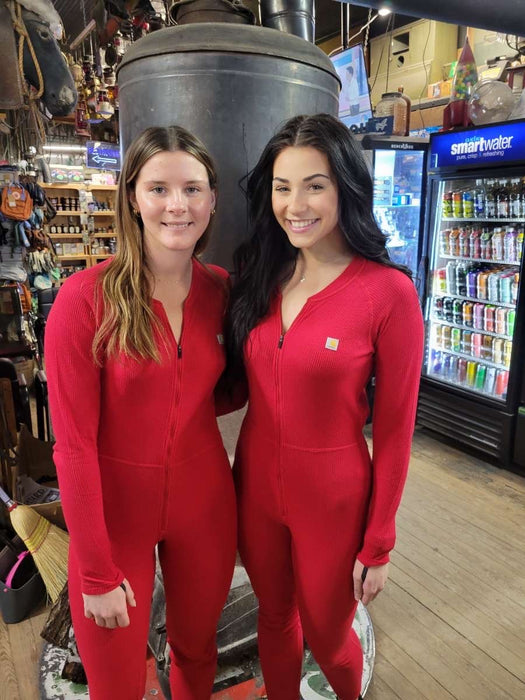 Two women smiling and wearing matching red Carhartt union suits, standing in a store with various items on the shelves in the background.