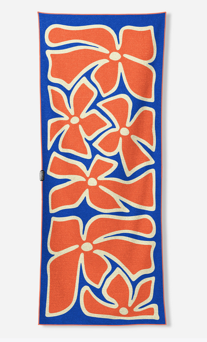 A rectangular beach towel with a bold pattern of orange stylized flowers outlined in cream on a royal blue background, shown flat against a light gray studio backdrop.